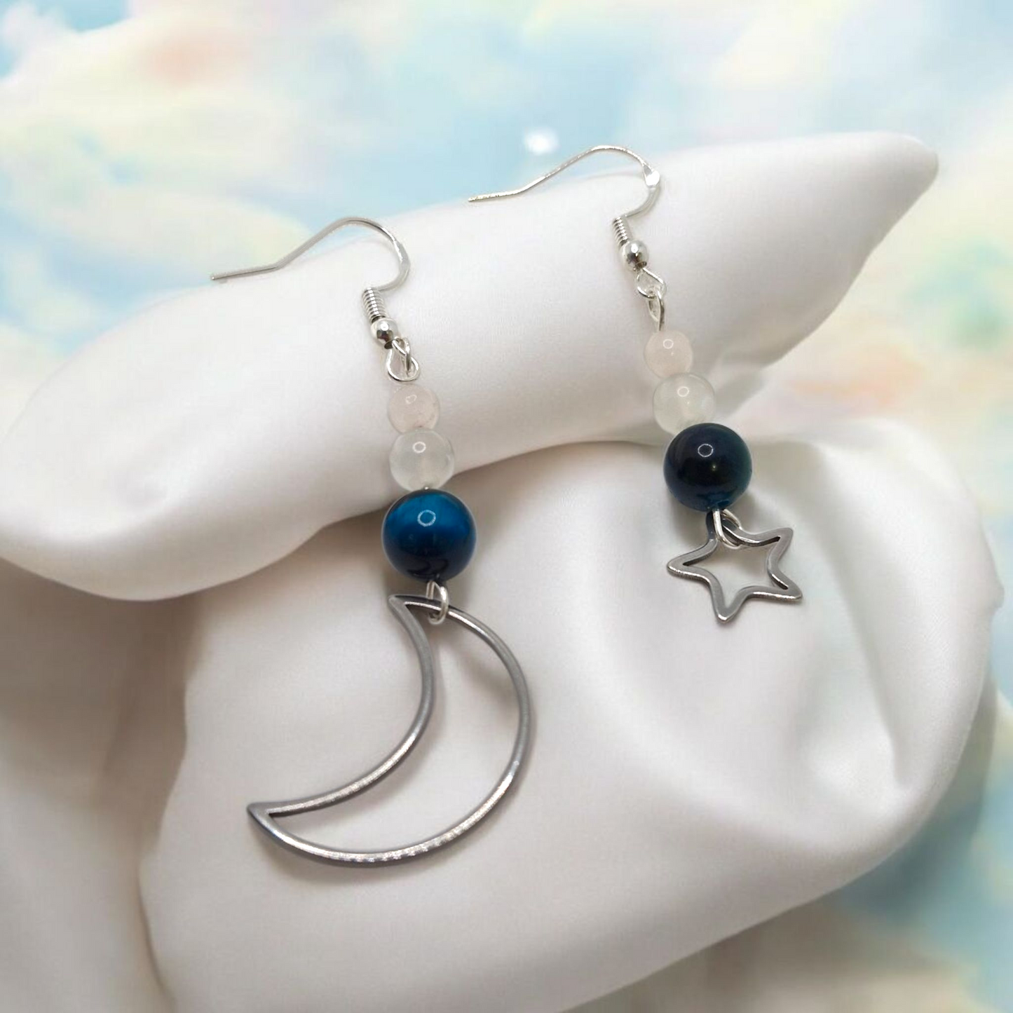 Earrings with moon, star, and blue bead designs on a white shell with a colorful background.