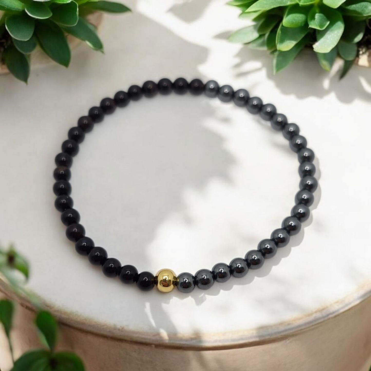 Black beaded bracelet with a gold accent on a white surface with green plants in the background