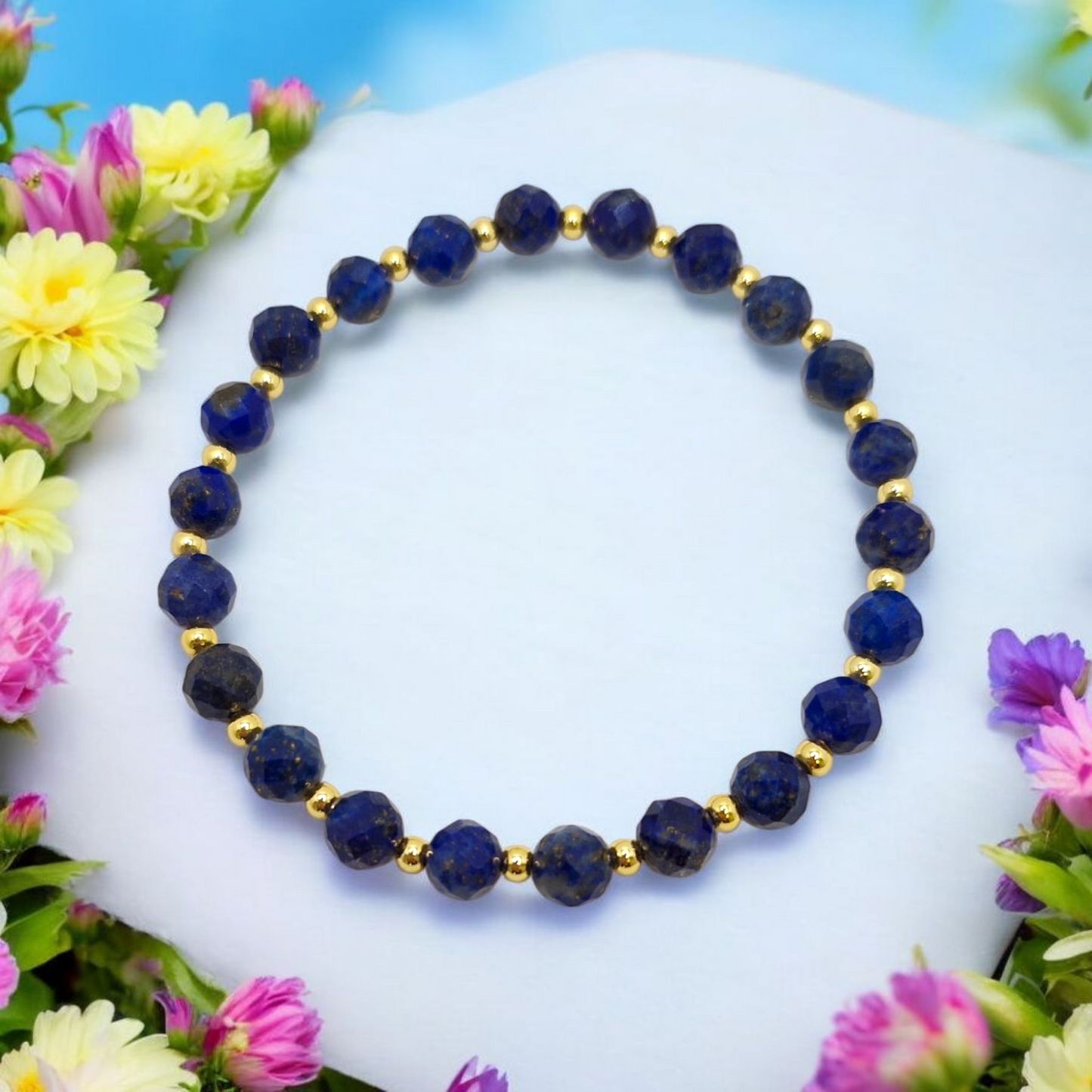 Blue beaded bracelet with gold accents on a white surface surrounded by colorful flowers