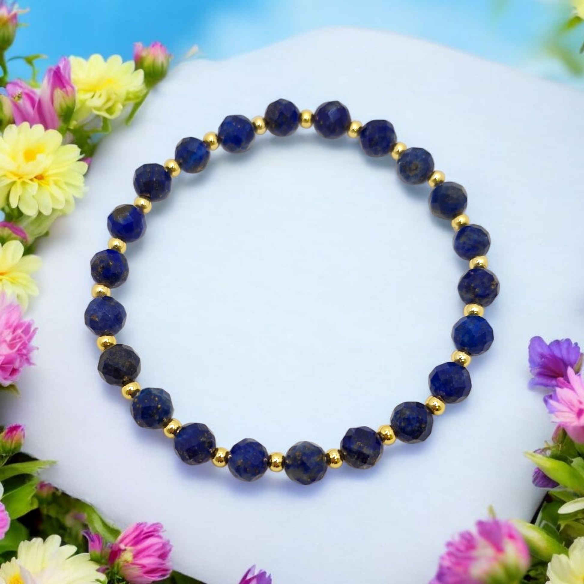 Blue beaded bracelet with gold accents on a white surface surrounded by colorful flowers