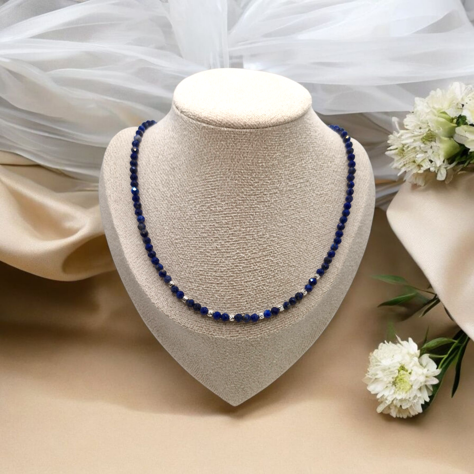Blue beaded necklace on a beige jewelry display with white flowers