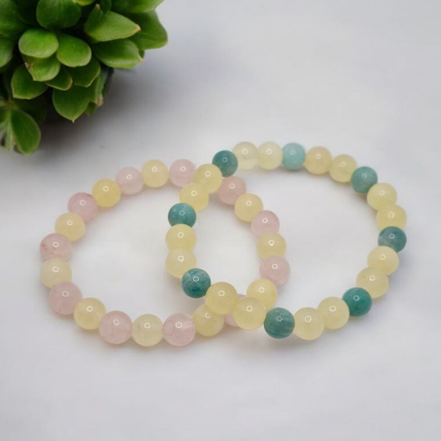 Two beaded bracelets in pastel colors on a white surface with a green plant in the corner.