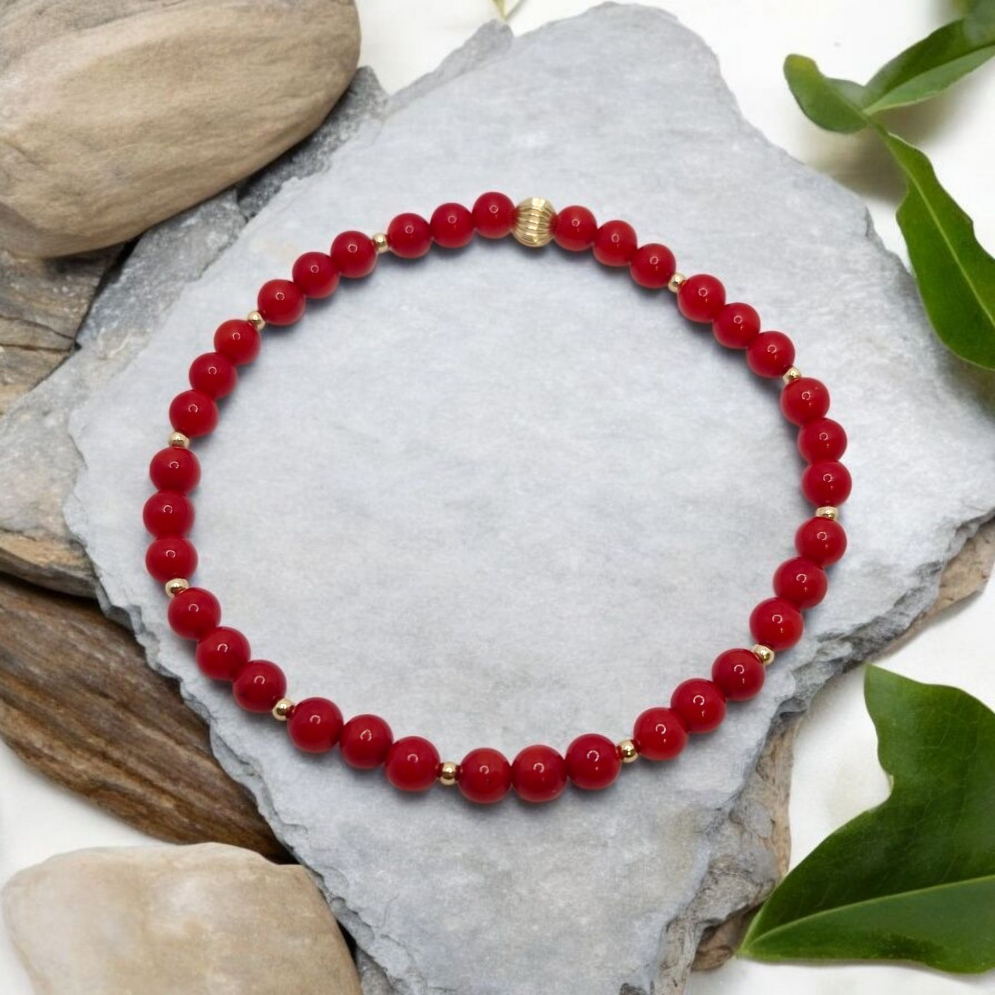 Red beaded bracelet with gold accents on a gray stone surface with green leaves.