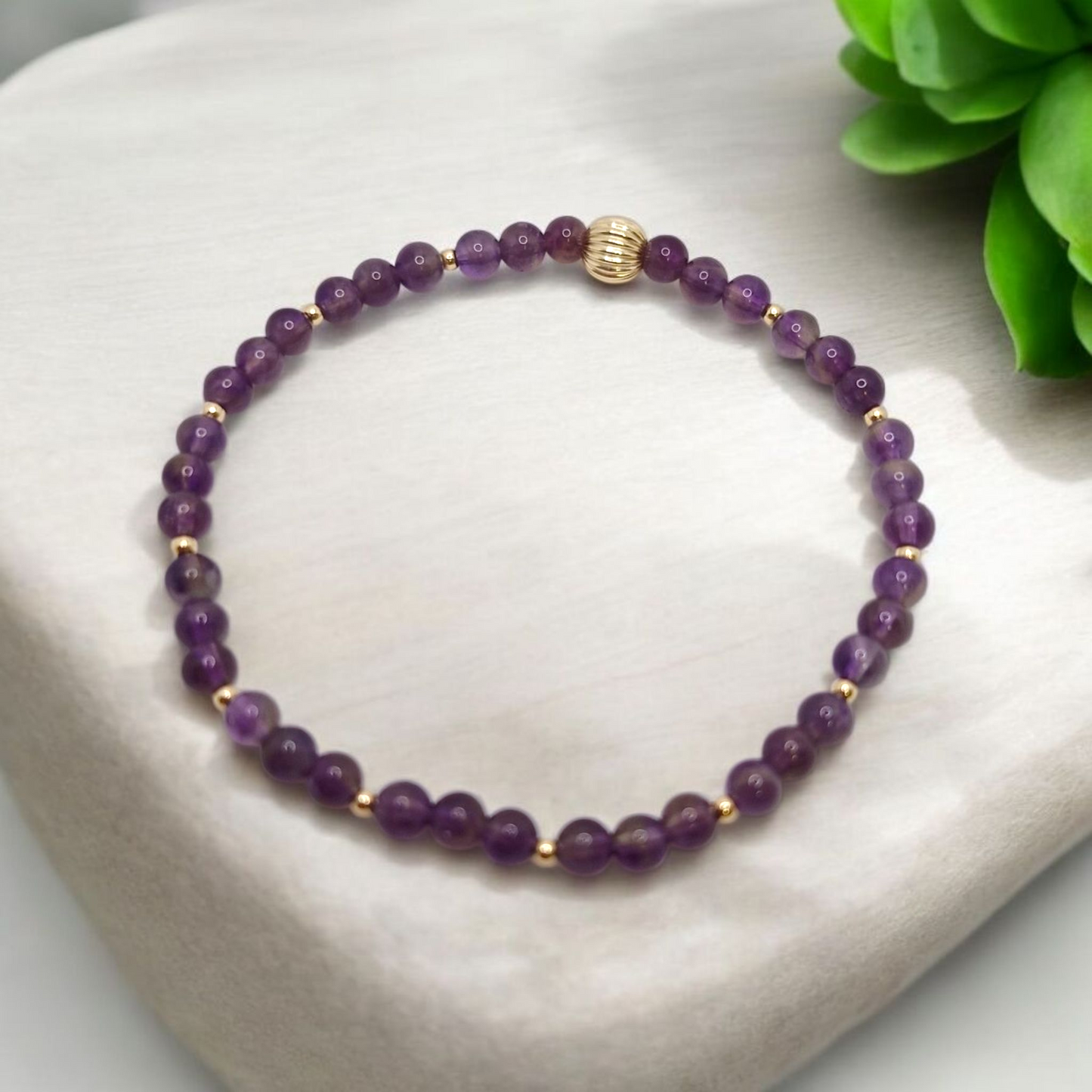 Purple beaded bracelet with gold accents on a white surface with a green plant in the background