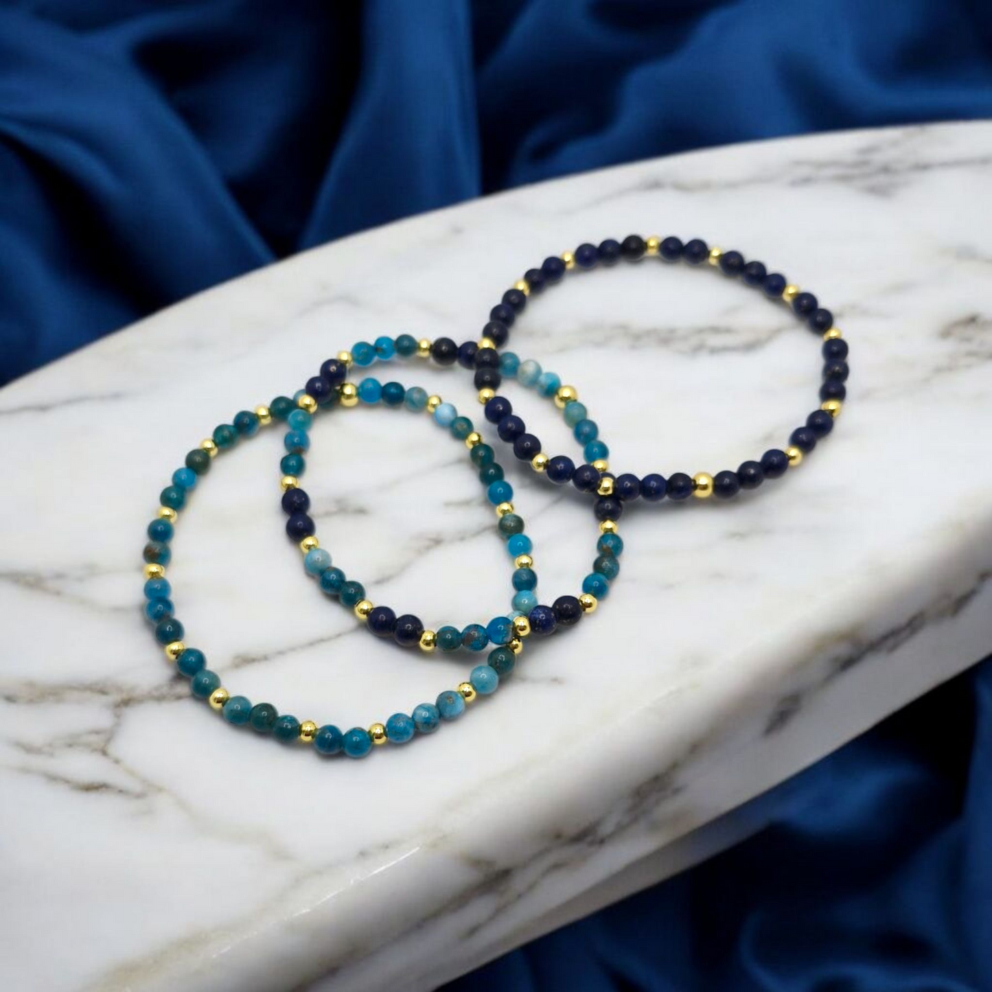 Two beaded bracelets on a marble surface with a blue fabric background