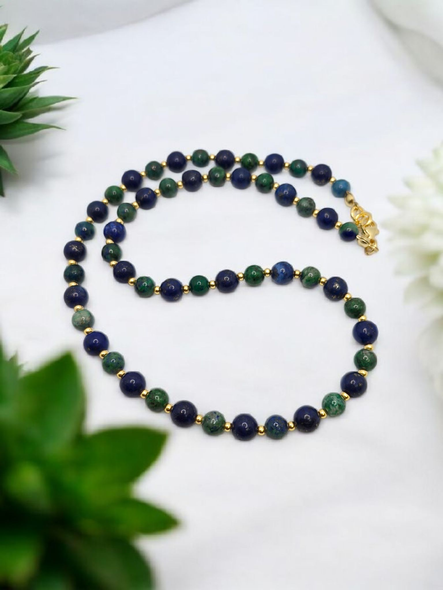Necklace with blue and green beads on a white surface with plants in the background