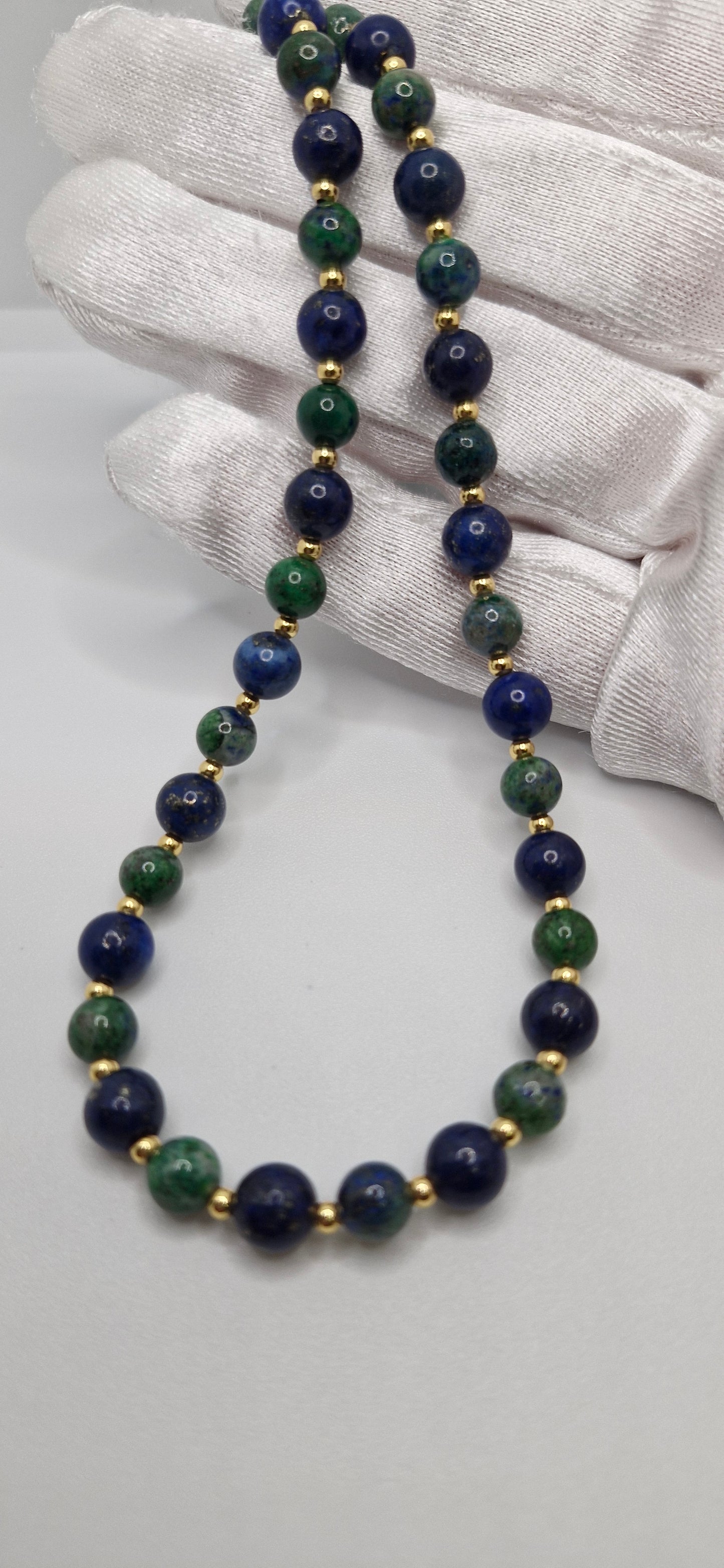 Necklace with blue and green beads held by a gloved hand on a light gray background